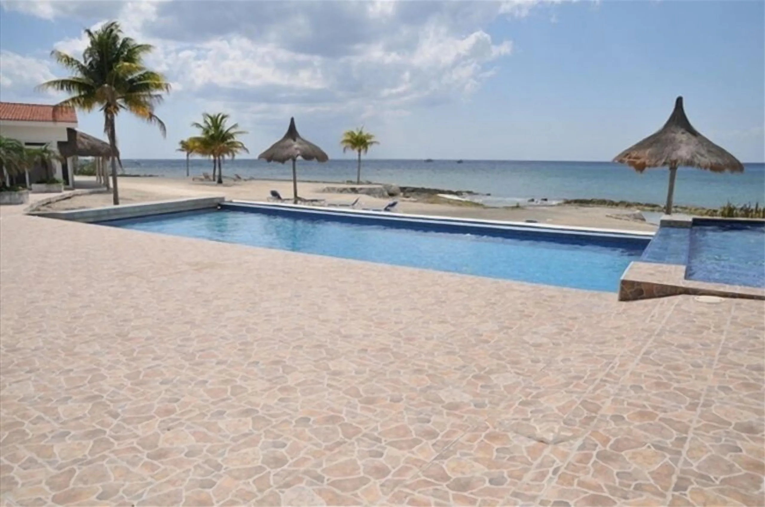 View across the pool deck to the beach, palm trees and thatched palapa umbrellas at Costa del Sol, Cozumel